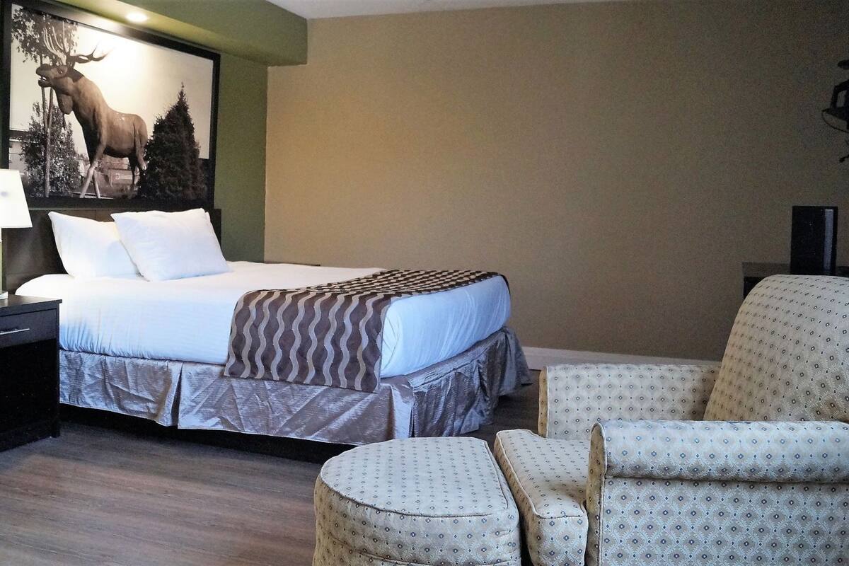 Quarto de hotel com cama, mesa de cabeceira, luminária, cadeira e televisão fixada na parede.