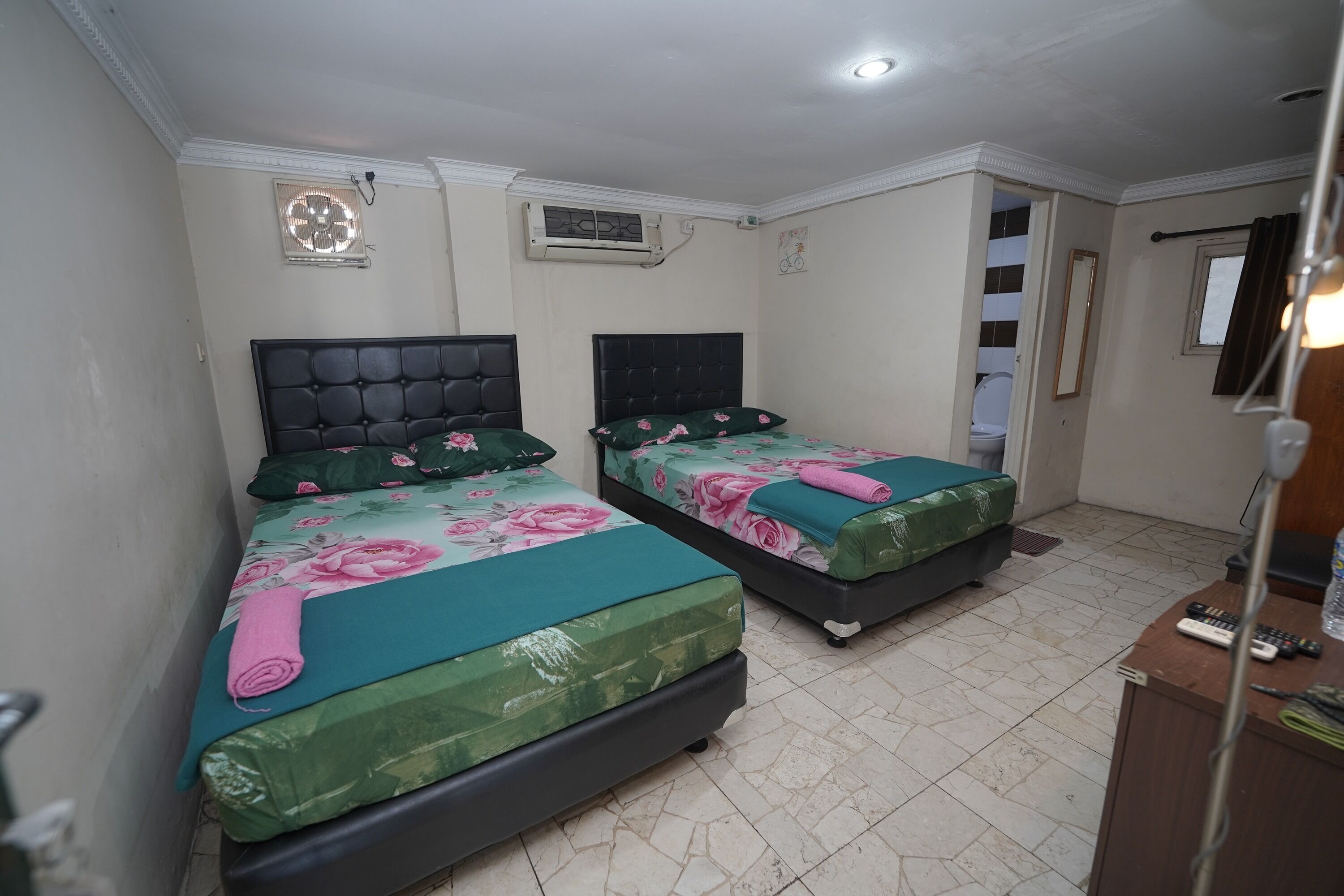 Une chambre avec deux lits, chacun agrémenté d’une couvre-lit à motifs floraux et d’une tête de lit. On y trouve deux climatiseurs fixés au mur, une petite fenêtre décorée d’un motif de vélo et un miroir également fixé au mur.