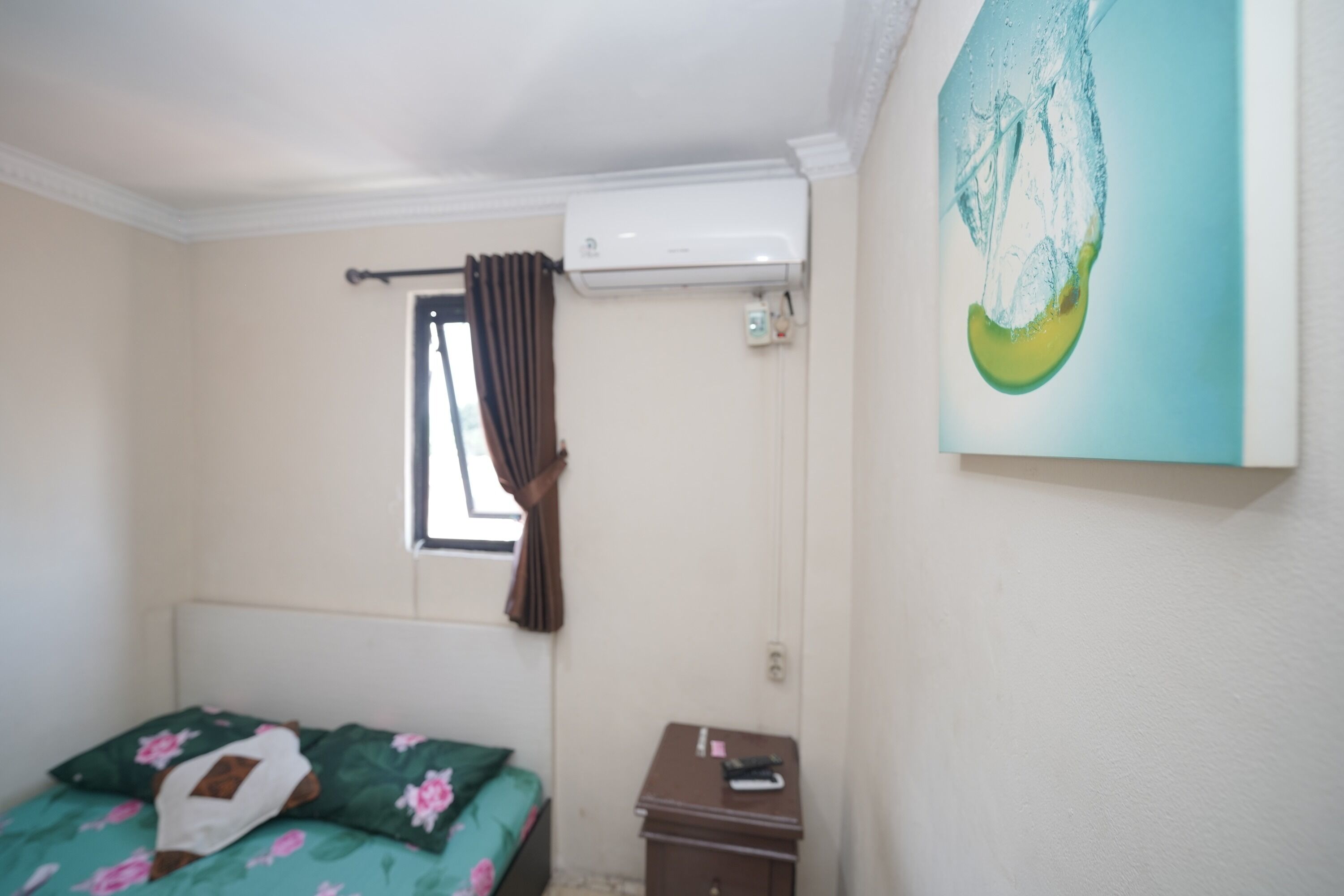 A bedroom with a bed, a nightstand, a window with curtains, an air conditioning unit, and a painting on the wall.