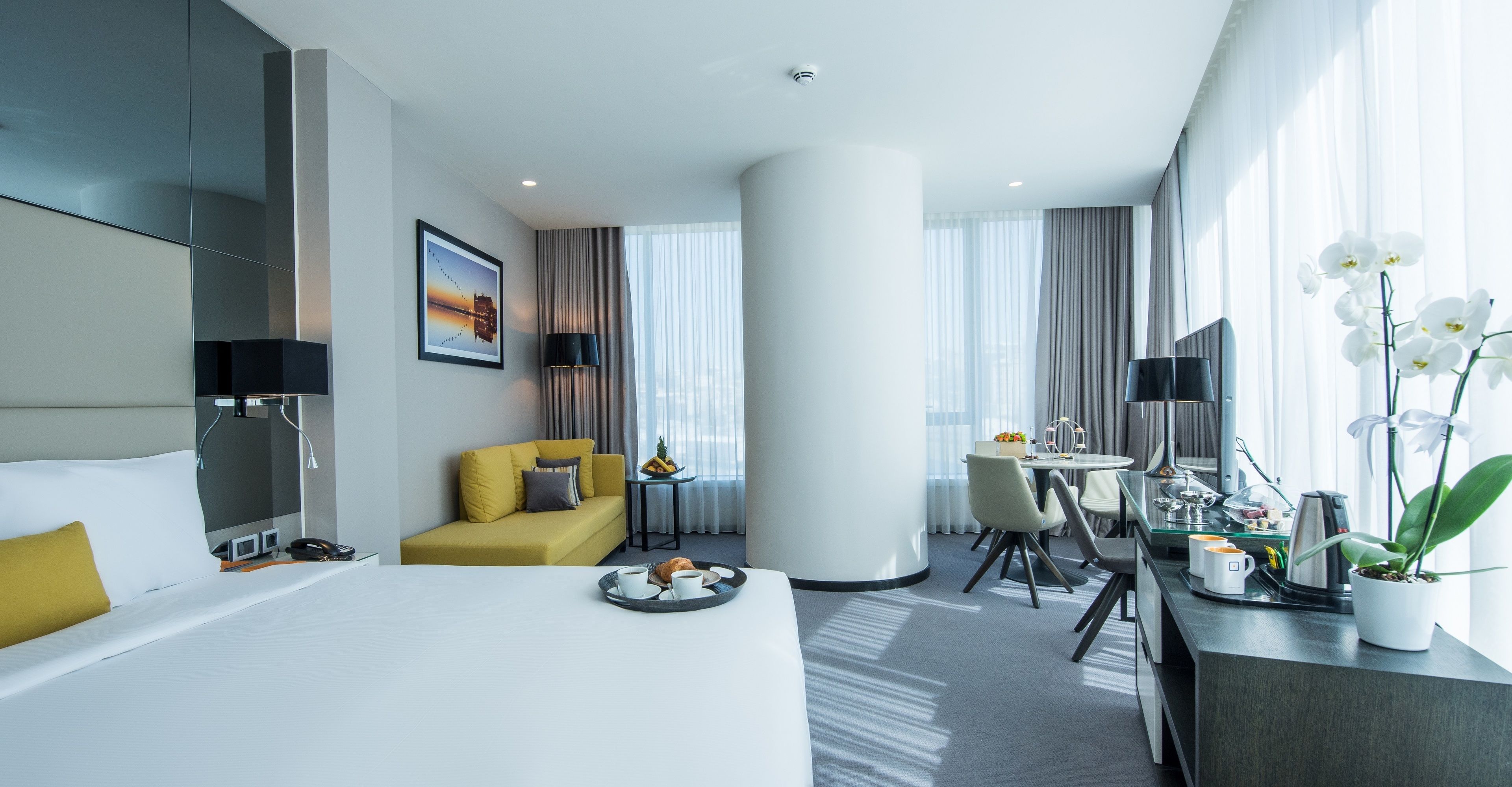 Una habitación de hotel moderna con una cama, un sofá amarillo, una zona de comedor y un gran ventanal con cortinas.