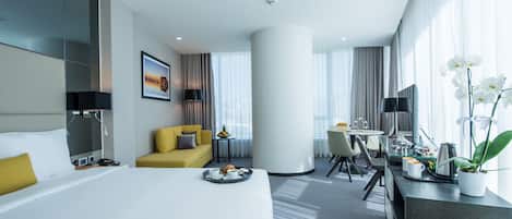 Una habitación de hotel moderna con una cama, un sofá amarillo, una zona de comedor y un gran ventanal con cortinas.