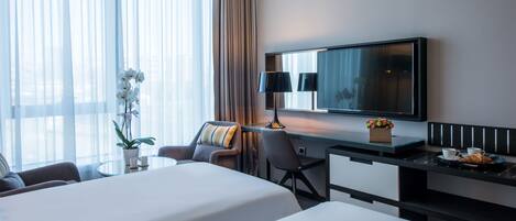 Ein Hotelzimmer mit zwei Betten, einem Schreibtisch mit Lampe, einem Sessel, einem kleinen Tisch mit Pflanze und einem an der Wand befestigten Fernseher.