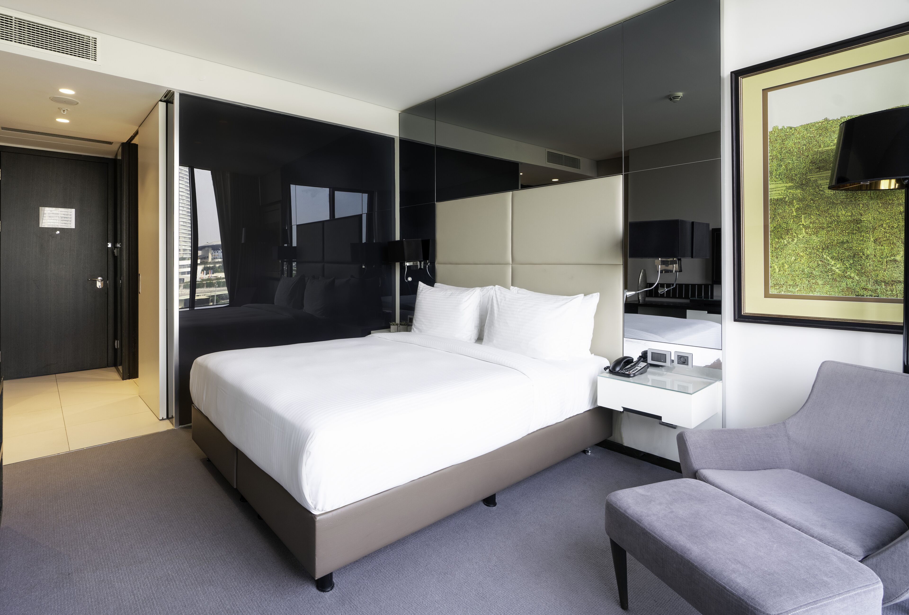 A modern hotel room with a large bed, a bedside table with a phone, a grey armchair, and a large mirror.