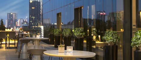 An outdoor seating area with tables and chairs, a potted plant, and a glass building in the background.