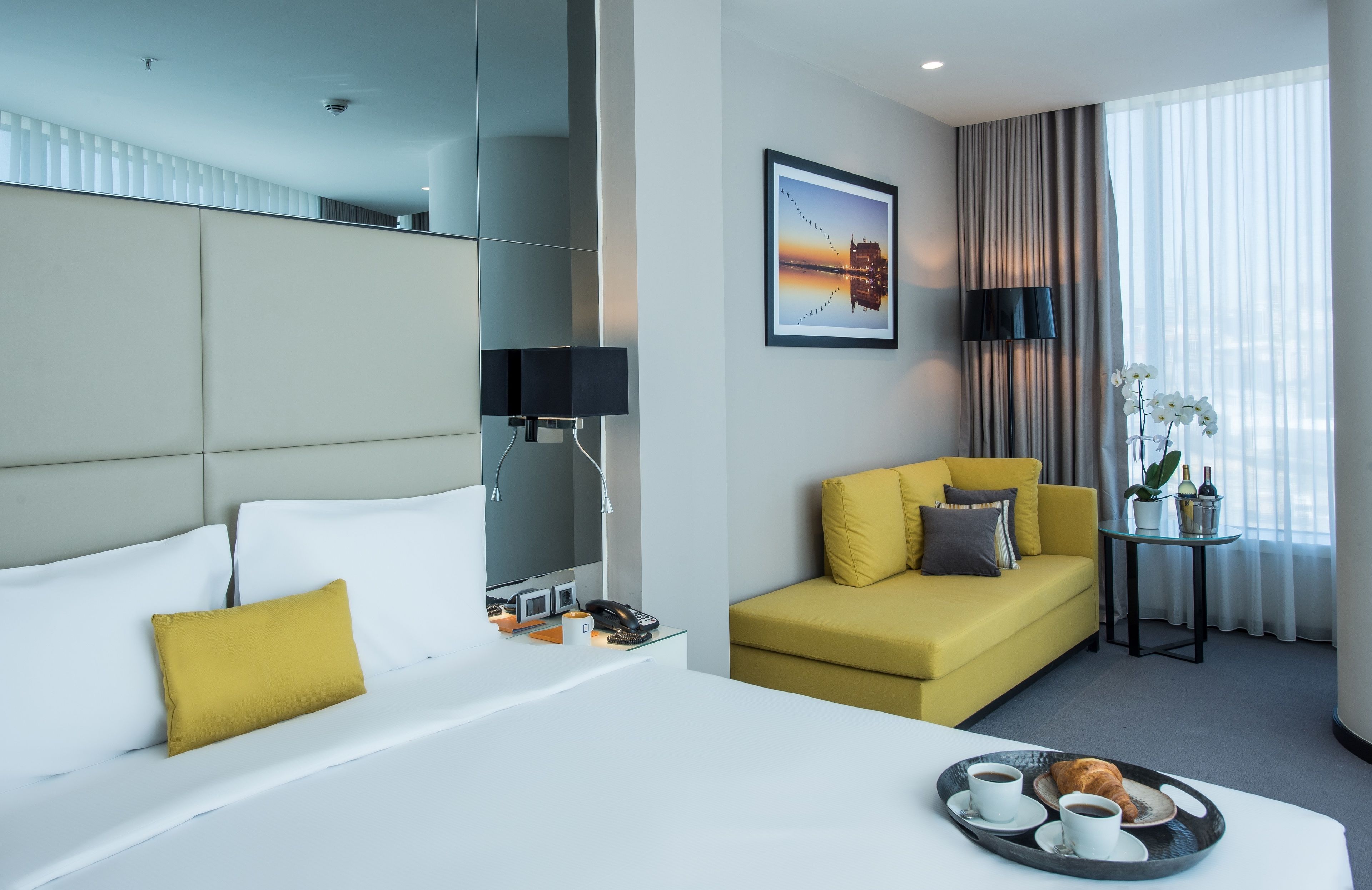 Habitación de hotel con cama, sofá, una mesita con una bandeja de comida, lámpara y ventana con cortinas.