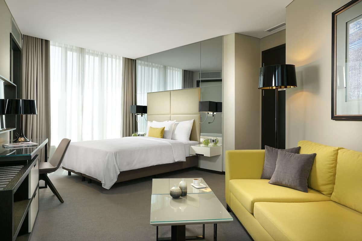 Ett hotellrum med en säng, ett skrivbord, en gul soffa, ett soffbord i glas och en stor spegel.