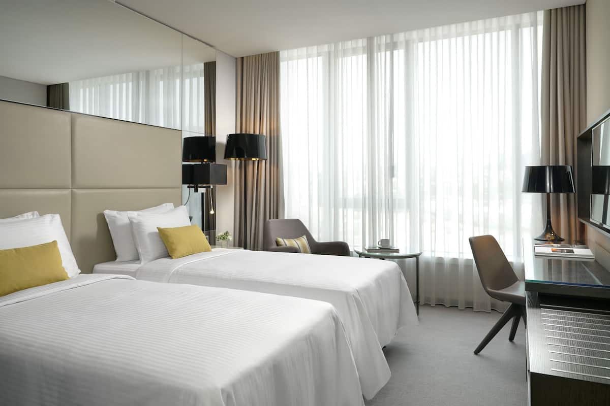 Ett hotellrum med två sängar, ett skrivbord, en stol, en tv och ett stort fönster med gardiner.