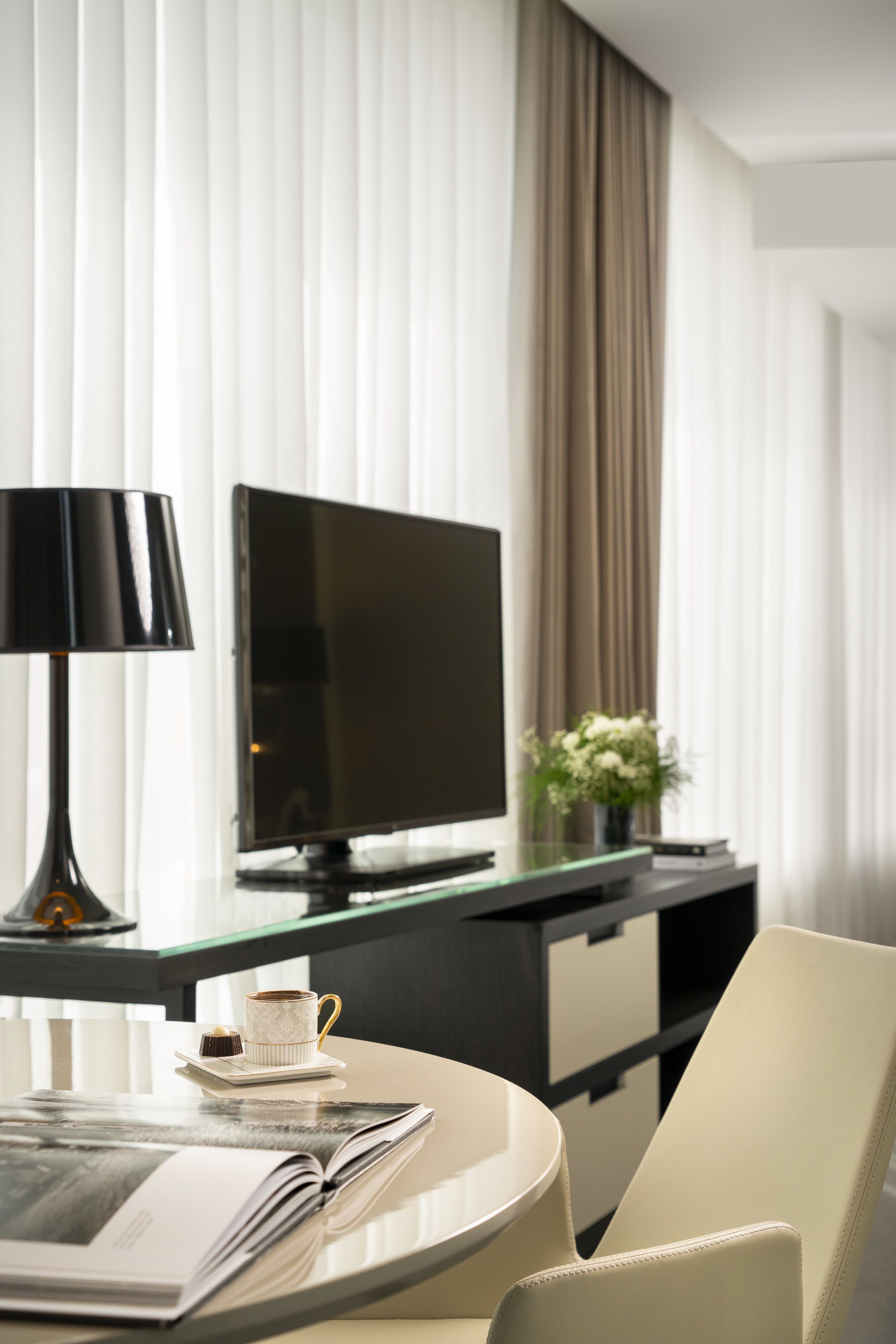 A modern hotel room with a round table, a television, a lamp, a cup, and a book.