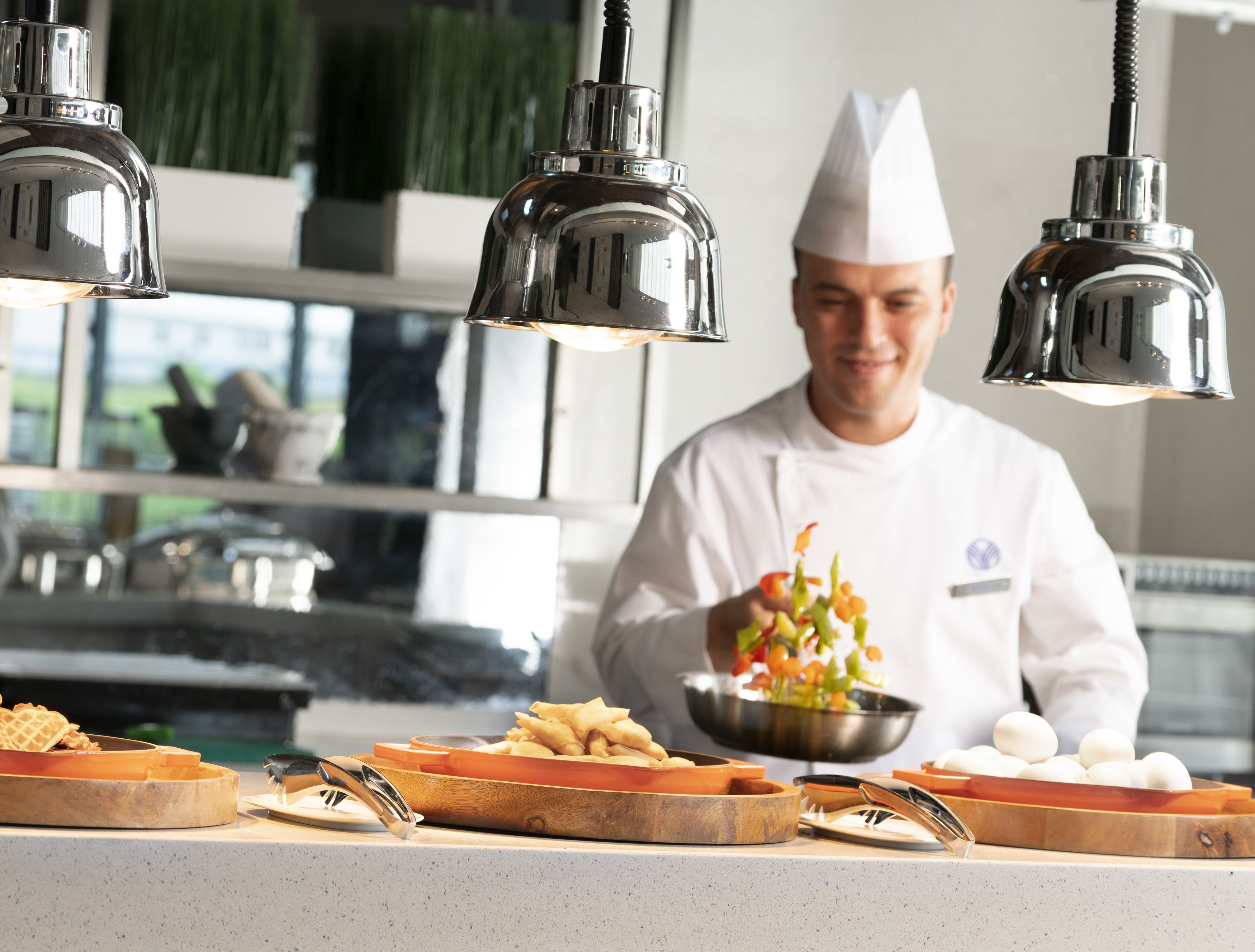 Un chef, vêtu d’un uniforme blanc avec une étiquette nominative, tient un saladier rempli de légumes. On peut voir différents plats sur le comptoir, notamment des frites et des œufs. La cuisine est dotée de luminaires suspendus et présente un design moderne.