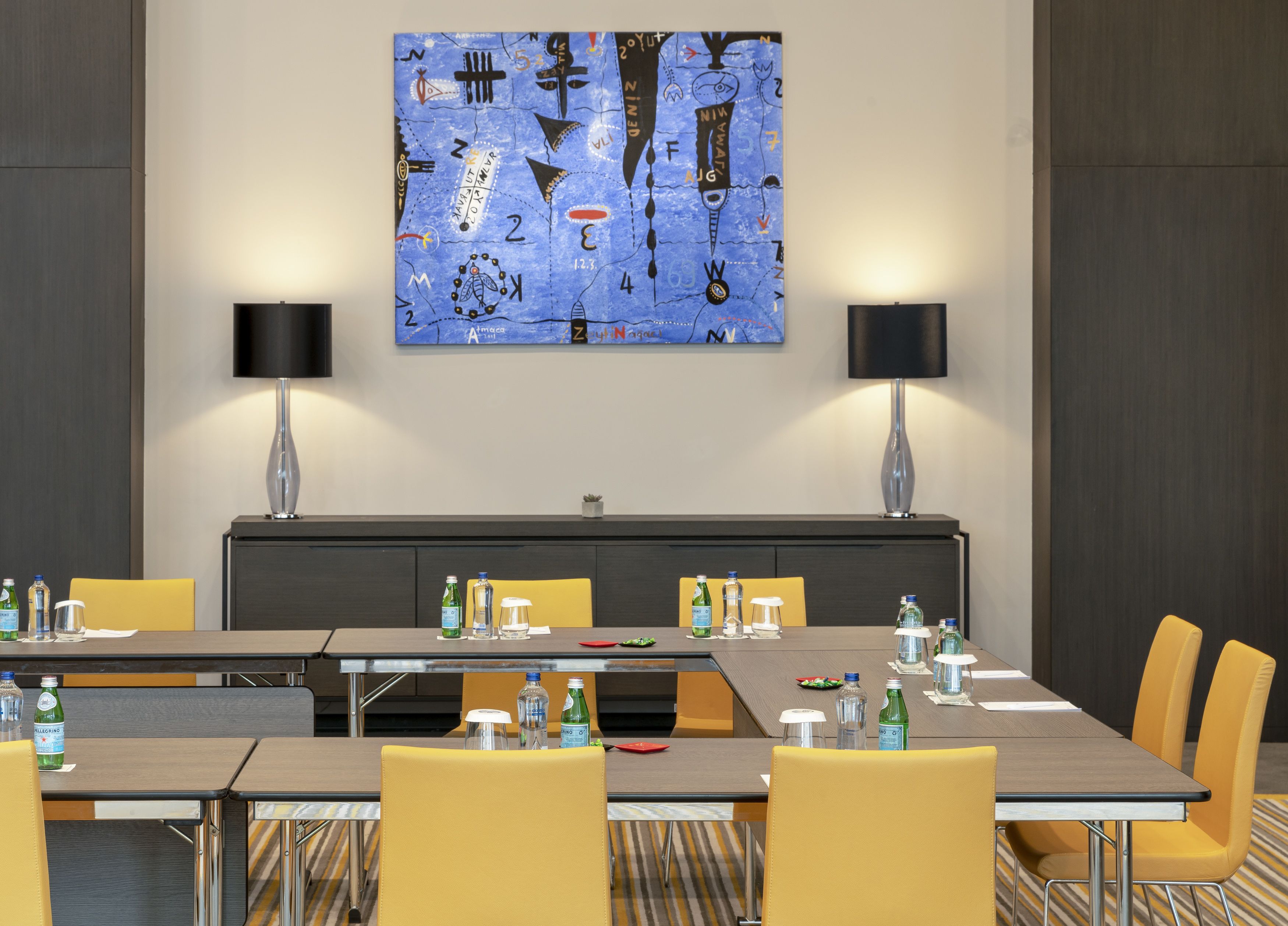 A conference room with a long table, yellow chairs, two lamps, and a large abstract painting on the wall.