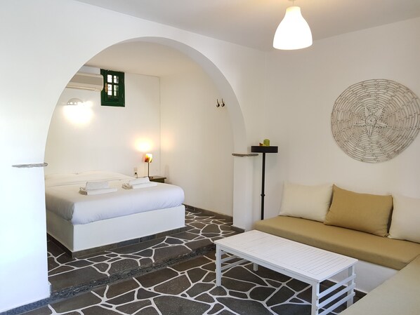 A hotel room with a bed, a sofa, a coffee table, a wall-mounted lamp, a decorative wall piece, and a tiled floor. - Andromeda (Sifnos)