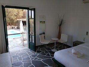 A hotel room with a bed, a small table, two chairs, a potted plant, and a view of an outdoor pool area. - Andromeda (Sifnos)