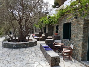 A stone-walled outdoor seating area with built-in benches and tables, surrounded by trees and a stone pathway. - Andromeda (Sifnos)