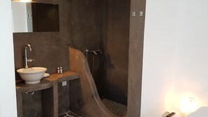 Une salle de bain avec un sol en pierre, un lavabo blanc, un miroir et un espace douche.