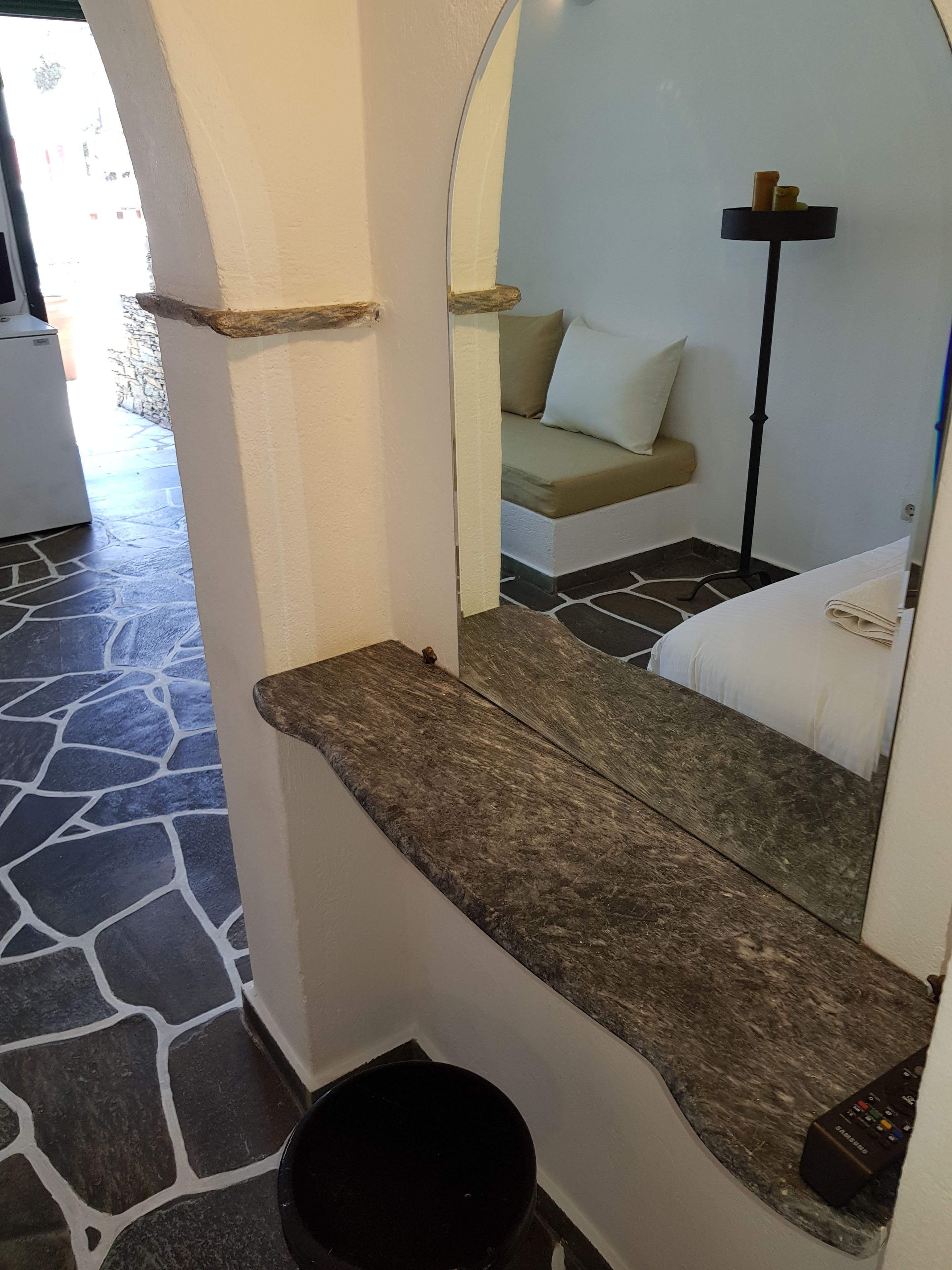 A room with a stone-tiled floor, a stone bench, a bed with white bedding, a bedside table, a lamp, and a remote control on the bed.