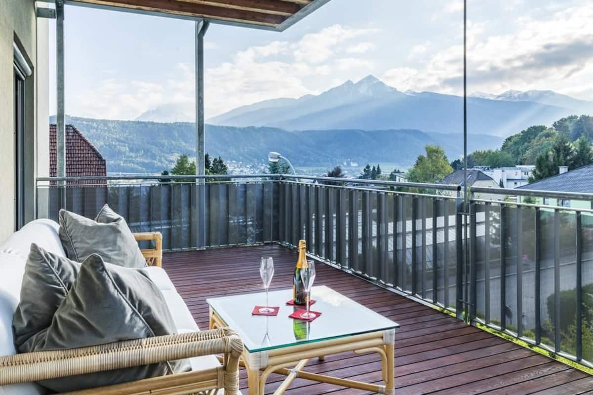 Un balcon avec un canapé en osier, une table en verre avec une bouteille et des verres, et une vue sur les montagnes.