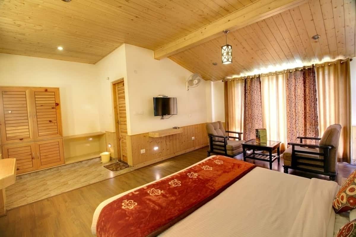 A hotel room with a wooden ceiling, a bed with a red bedspread, two chairs, a small table, a TV mounted on the wall, and a window with patterned curtains.