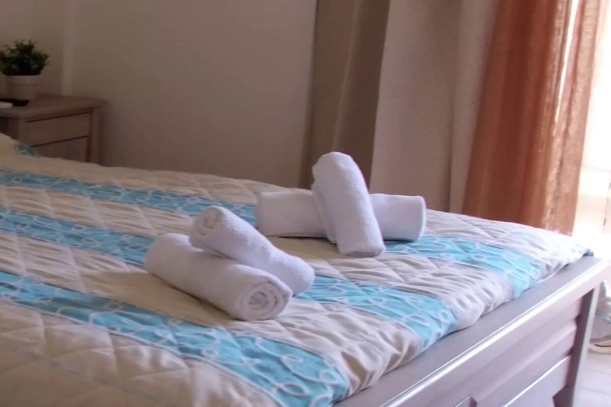 Ein ordentlich gemachtes Bett mit einer blau-weißen Bettdecke, zwei zusammengerollten Handtüchern und einem kleinen Nachttisch mit einer Pflanze.