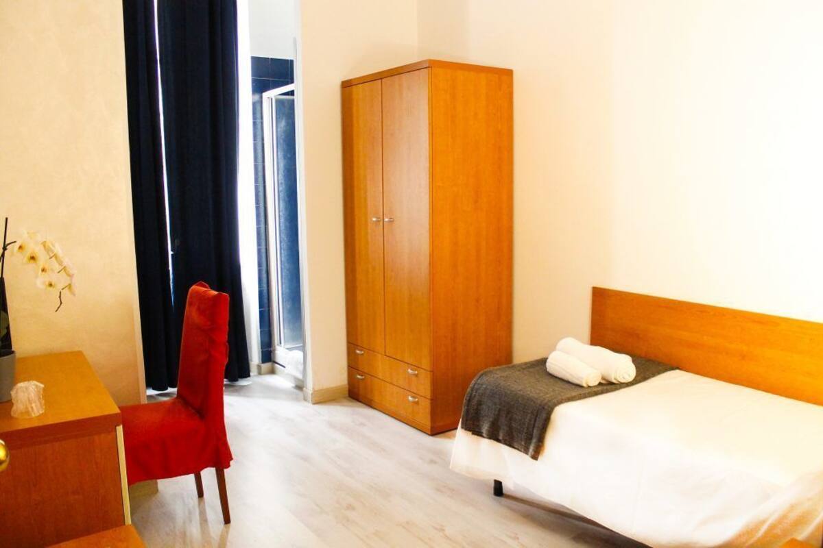 A hotel room with a bed, a wooden wardrobe, a desk with a chair, and a view of a corridor.