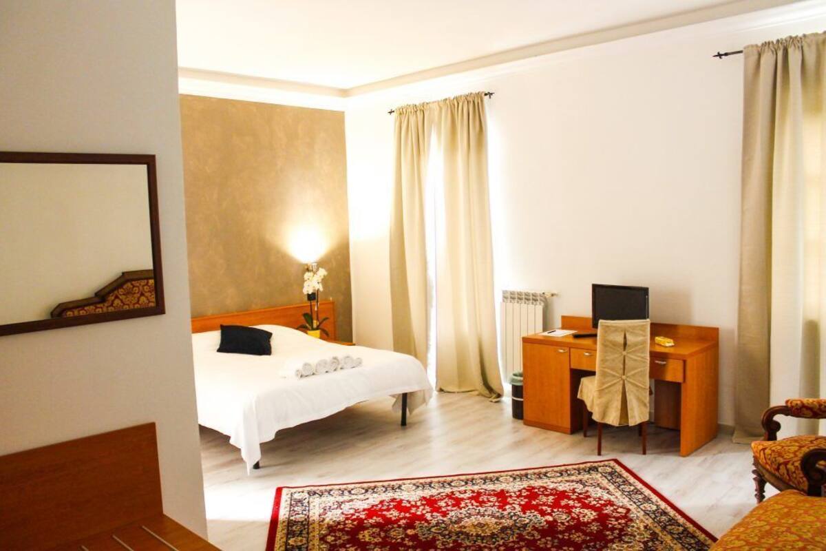 A hotel room with a bed, a desk, a chair, a mirror, and a red patterned rug.