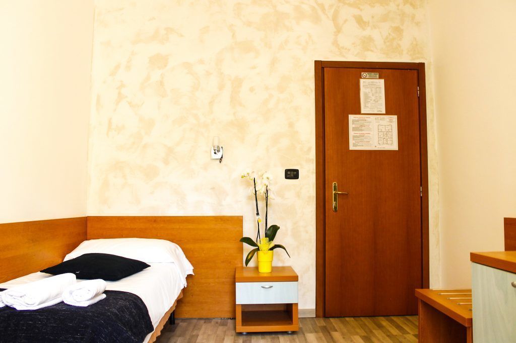 A small, neatly arranged bedroom with a wooden bed, a nightstand with a vase of flowers, a wooden wardrobe, and a door with a notice on it.