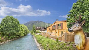 A wooden cabin with a river in the background and a deer statue in the foreground. - The Kaprun Edition (Kaprun)