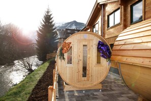 Una sauna in legno con una porta, circondata da un pontile in legno, vicino a un fiume e montagne.