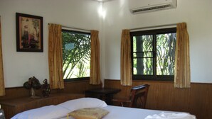 Habitación de hotel con una cama grande, dos ventanas con cortinas, un escritorio de madera, una silla y un aire acondicionado instalado en la pared.