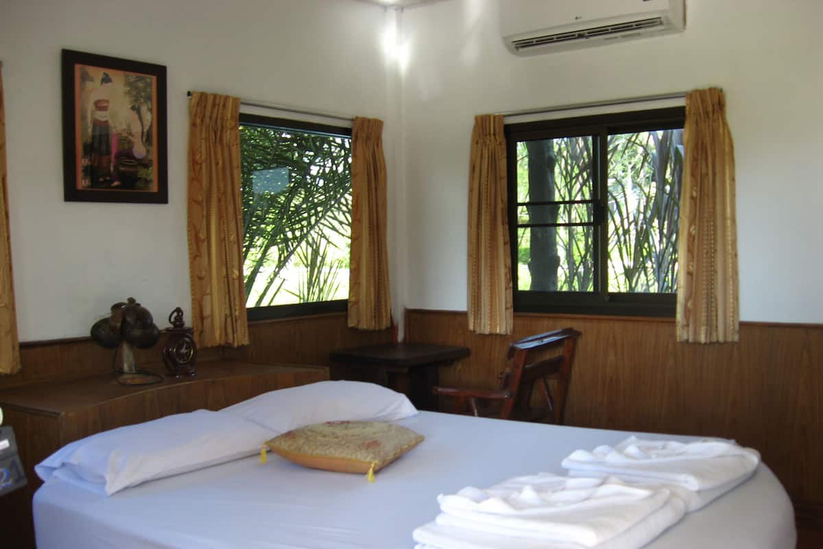 A hotel room with a large bed, two windows with curtains, a wooden desk, a chair, and a wall-mounted air conditioner.