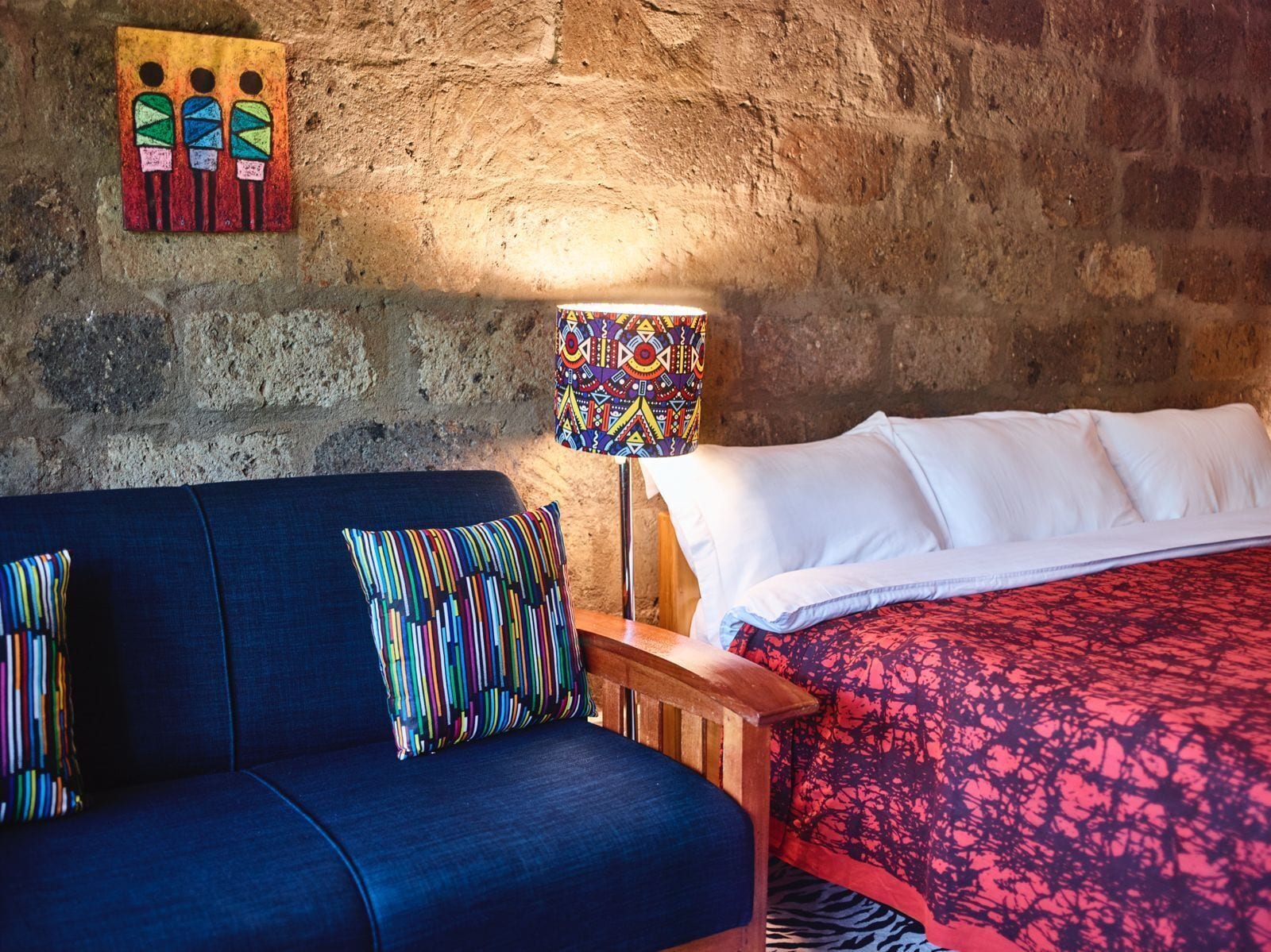 Una habitación con un sofá azul, una mesita de noche de madera con una lámpara y una cama con ropa de cama estampada.