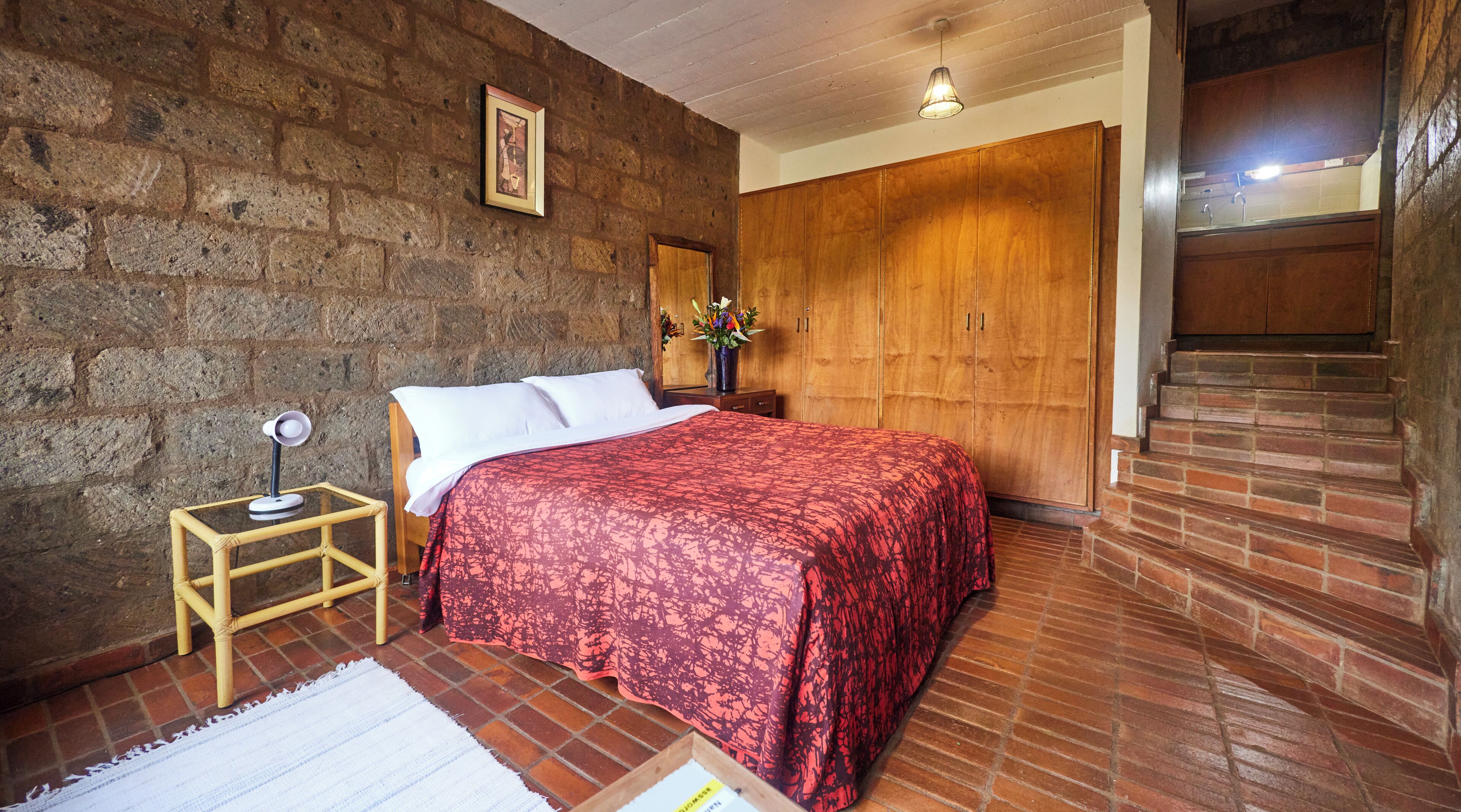 A bedroom with a brick wall, a wooden wardrobe, a bed with a red patterned bedspread, a nightstand with a lamp, and a small table with a vase of flowers.