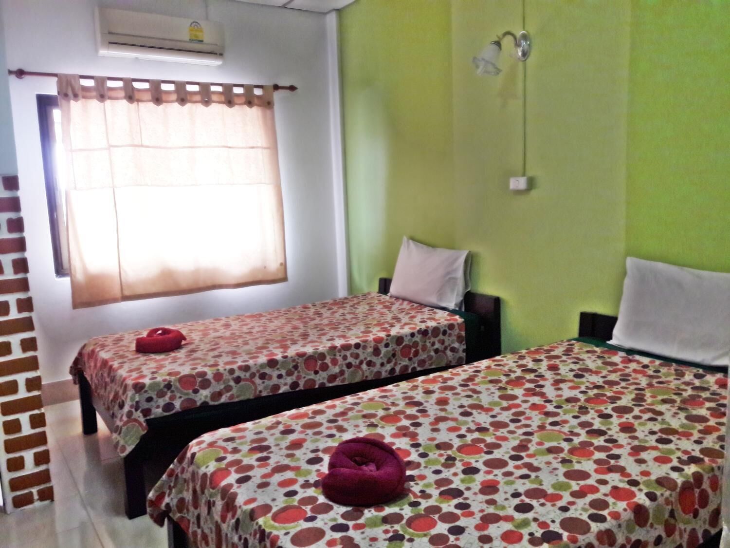 A room with two beds, each with a floral-patterned bedspread and a red heart-shaped pillow. There is a window with a curtain, a wall-mounted air conditioner, and a wall-mounted light fixture.