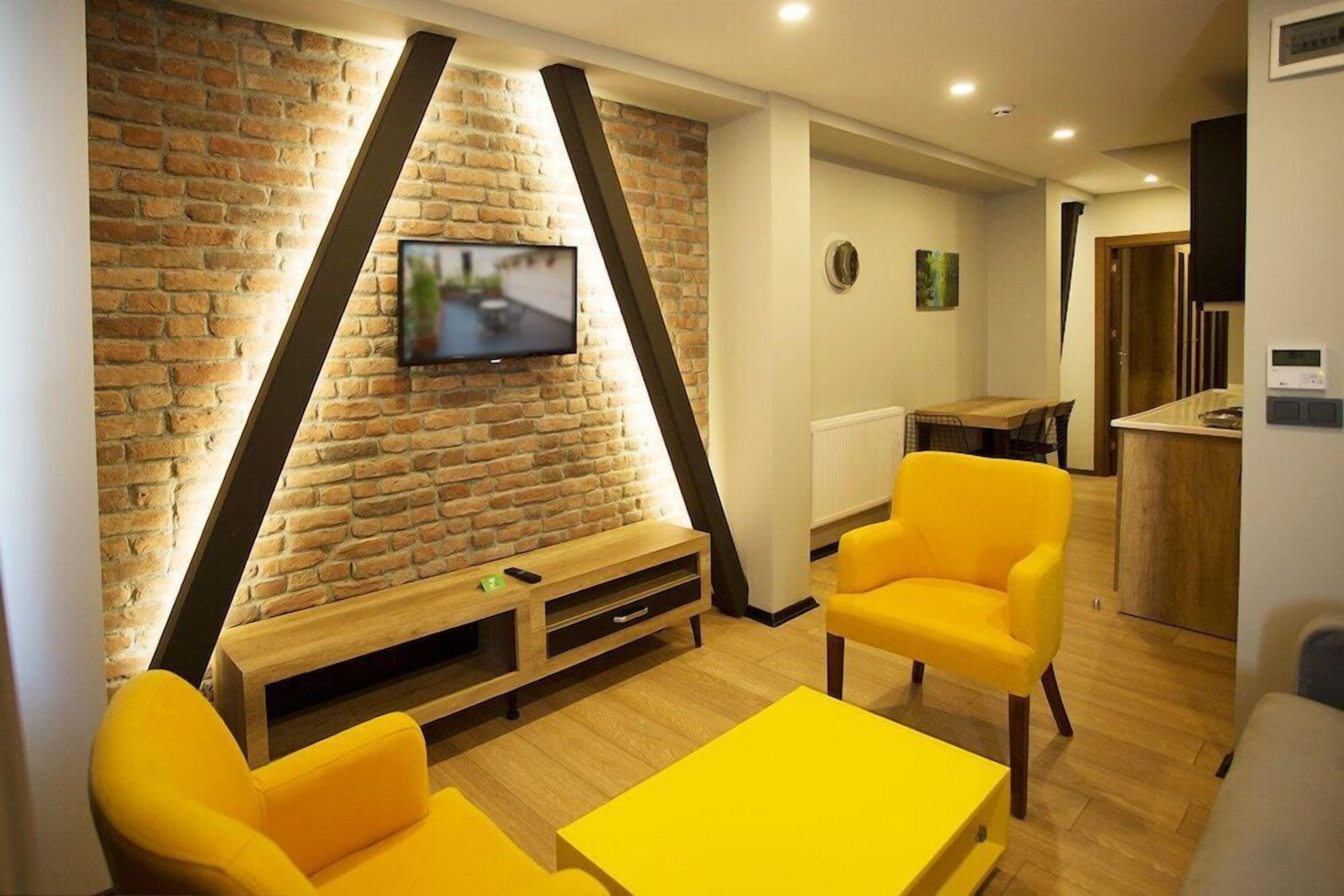 Una sala de estar moderna con pared de ladrillo, televisor de pantalla plana, mobiliario amarillo y una mesa de centro de madera.