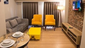 A modern living room with a sofa, two yellow armchairs, a coffee table, a TV mounted on a brick wall, and a dining area with a table set for two. - Long Stay İstanbul (Taksim - Beyoglu - Istanbul)