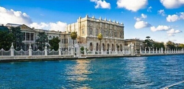 A large, historic building with a symmetrical facade, arched windows, and a central tower, situated along a waterfront with a white fence and palm trees. - Long Stay İstanbul (Taksim - Beyoglu - Istanbul)