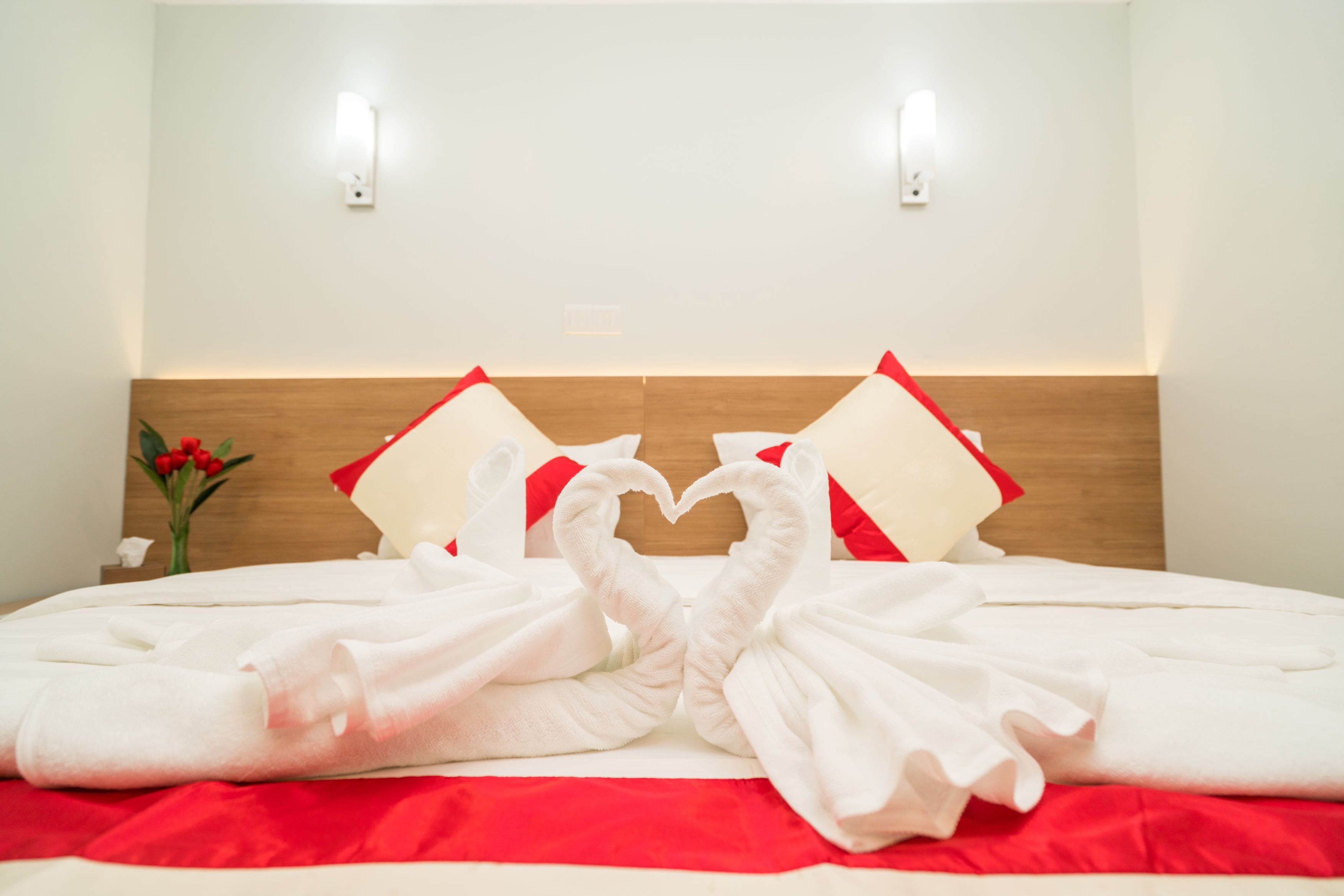 A hotel room with a neatly made bed, two pillows, a vase with flowers, and two wall-mounted lights.