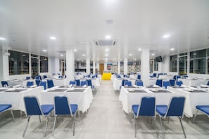 A large, well-lit conference room with rows of tables and chairs, a stage at the front, and large windows along the walls. - Sakura Hotel (Yangon)