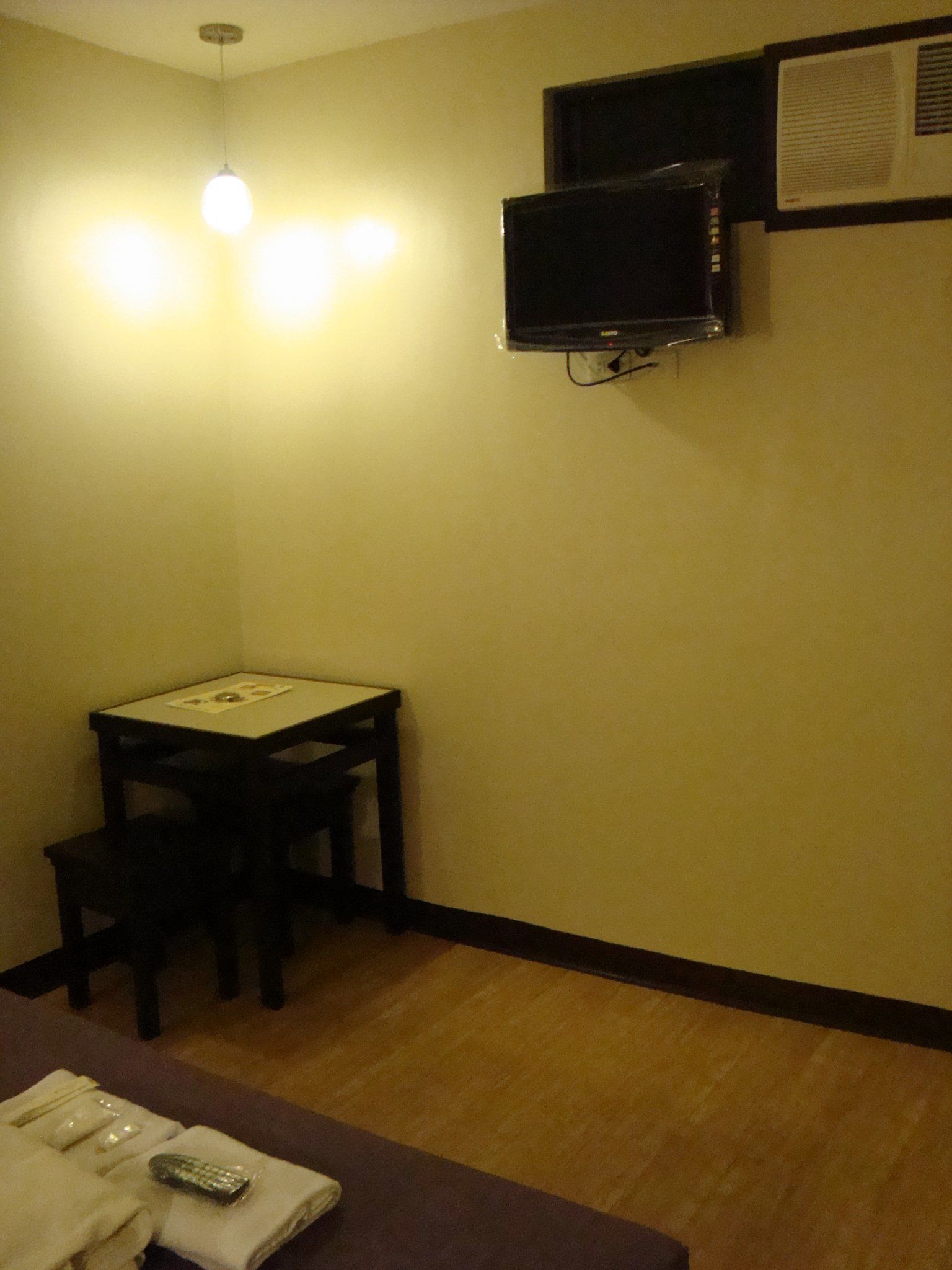 Una habitación pequeña con una mesa, una silla, un televisor montado en la pared y una lámpara en el techo.