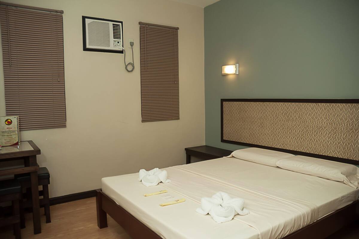 A hotel room with a bed, a desk with a chair, a telephone, and a wall-mounted air conditioner.