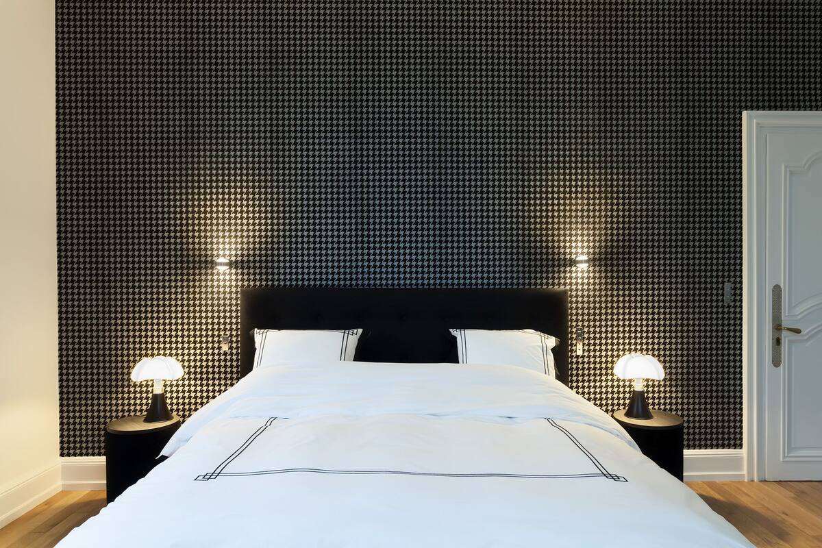 A neatly made bed with white linens and a black headboard, flanked by two bedside tables with lamps, set against a patterned wallpaper.