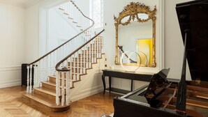 A grand piano, a staircase with white balusters, and a large ornate mirror in a spacious room with wooden flooring. - Villa Camoufle (Metz)