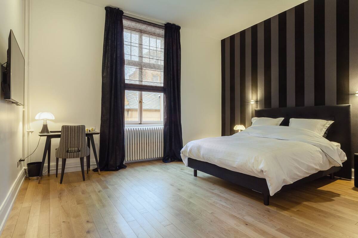 A bedroom with a bed, a chair, a table, a window with curtains, and a radiator.