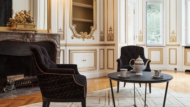 A luxurious room with a fireplace, ornate wall decorations, a black armchair, a round table with tea set, and a large mirror. - Villa Camoufle (Metz)