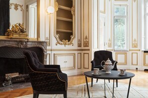 Une chambre luxueuse avec une cheminée, des décorations murales raffinées, un fauteuil noir, une table ronde avec un service à thé et un grand miroir.