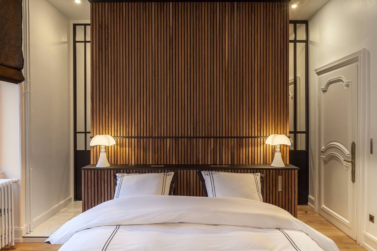 A hotel room with a large bed, two bedside lamps, a wooden panel headboard, and a classic door.