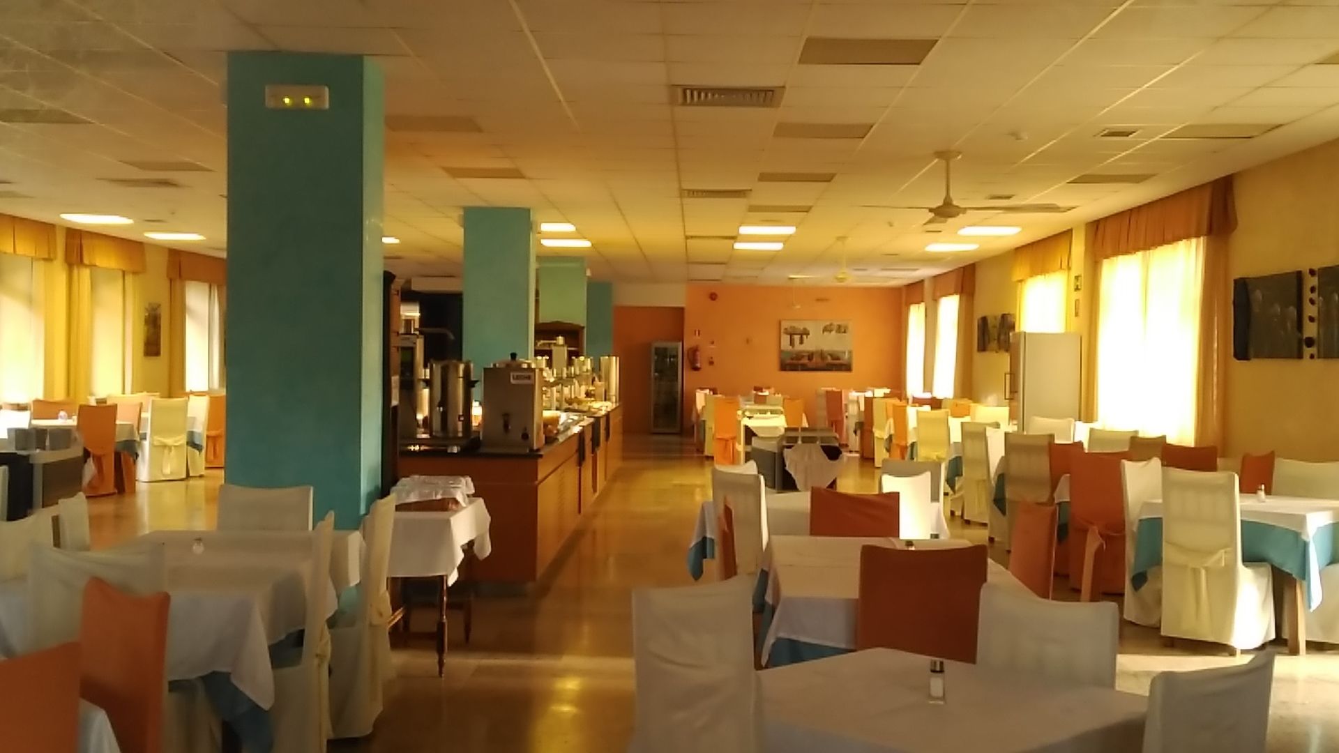 A spacious dining area with multiple tables and chairs, a central counter, and a kitchen in the background.