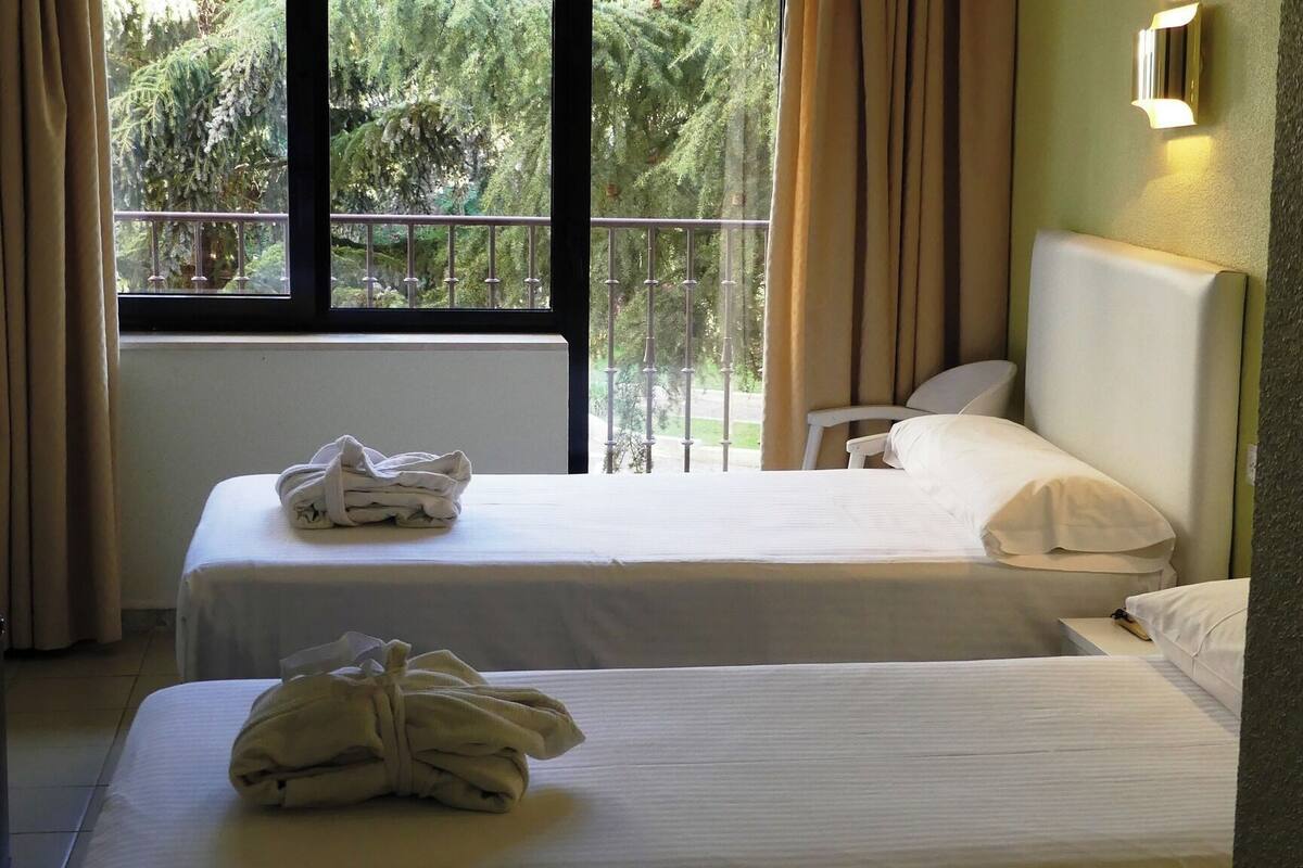 Een hotelkamer met twee bedden, een raam met gordijnen en uitzicht op bomen.
