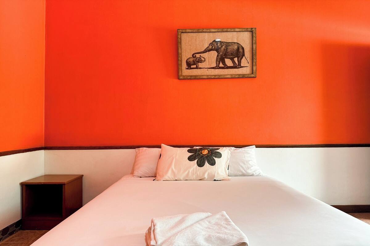 A hotel room with a large bed, orange walls, and a framed picture of elephants on the wall.