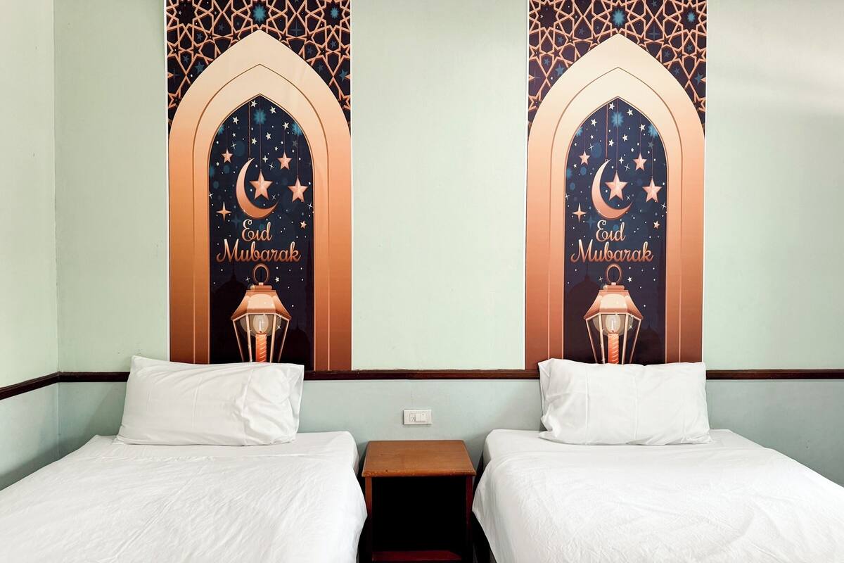 A hotel room with two beds, a nightstand, and a wall decorated with a moon and stars design, including the text "Eid Mubarak".