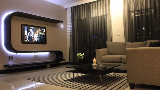 A modern living room with a flat-screen TV mounted on a curved wall, a sofa, a coffee table, and a decorative plant. - Yamsafer Homes 4th Circle (Amman)